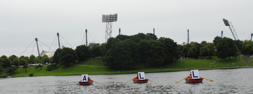 Photograph of three red rowing boats lined up on the Olympic lake. A white placard with a black capital letter is attached to the front of each boat; when read in sequence, they form the word "ALL". The silhouette of the Olympic Stadium can be seen in the background.