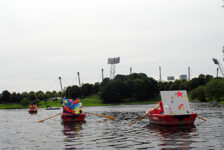 Photograph of three red rowing boats on the Olympic lake. A large canvas painted with different colourful star motifs is attached to each of the boats. The tent roof of the Olympic Stadium can be seen in the background.