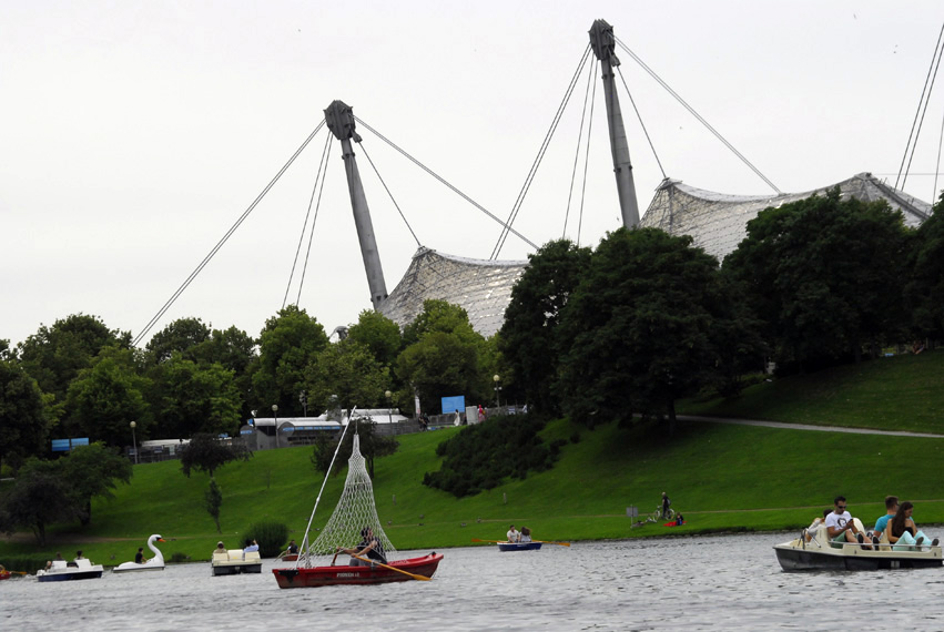 Fotografie des Olympiasees mit dem Zeltdach des Olympiastadions im Hintergrund. In der Mitte des Sees zu sehen ist ein rotes Ruderboot, das von einer Art Zeltdach aus weißem Gitternetz überspannt wird. Ein Ruderer sitzt unter dem Dach. Auf dem See sind zudem noch weitere Ruderboote unterwegs.
