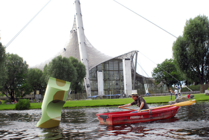 Photograph of a red rowing boat on the Olympic lake. The rower of the boat appears to be rowing around a larger-than-life green buoy.