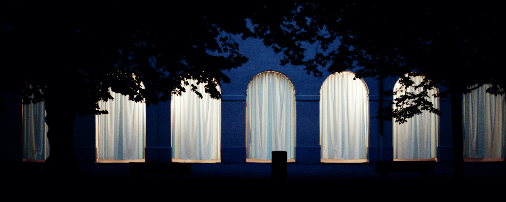 Frontale Ansicht der Hofgartenarkaden bei Nacht. Die Arkadenbögen sind mit weißen Vorhängen verhangen, die dahinterliegenden Arkadengänge sind erleuchtet und das Licht scheint durch die Vorhänge hindurch.