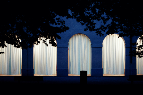 Frontale Ansicht der Hofgartenarkaden bei Nacht. Die Arkadenbögen sind mit weißen Vorhängen verhangen, die dahinterliegenden Arkadengänge sind erleuchtet und das Licht scheint durch die Vorhänge hindurch.
