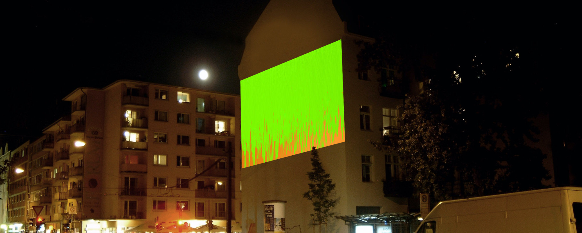 Photography of the large-scale projection "Bunter Abend" by Wolfgang Aichner and Thomas Huber on the facade of a building at Müllerstr. 10 at night. In the projection, two thick colours - green and red - flow over each other from the upper edge of the projection area.