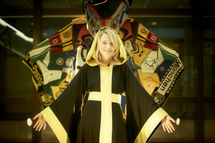 Photograph of the Münchner Kindl in the foreground, wearing the traditional black and yellow monk's habit, with arms outstretched. In the background behind him is another cloak based on the Münchner Kindl's robe. However, this cloak is made up of textile items from various Munich clubs, which have been assembled to form a patchwork 'Munich club collage' in the form of a Münchner Kindl robe.