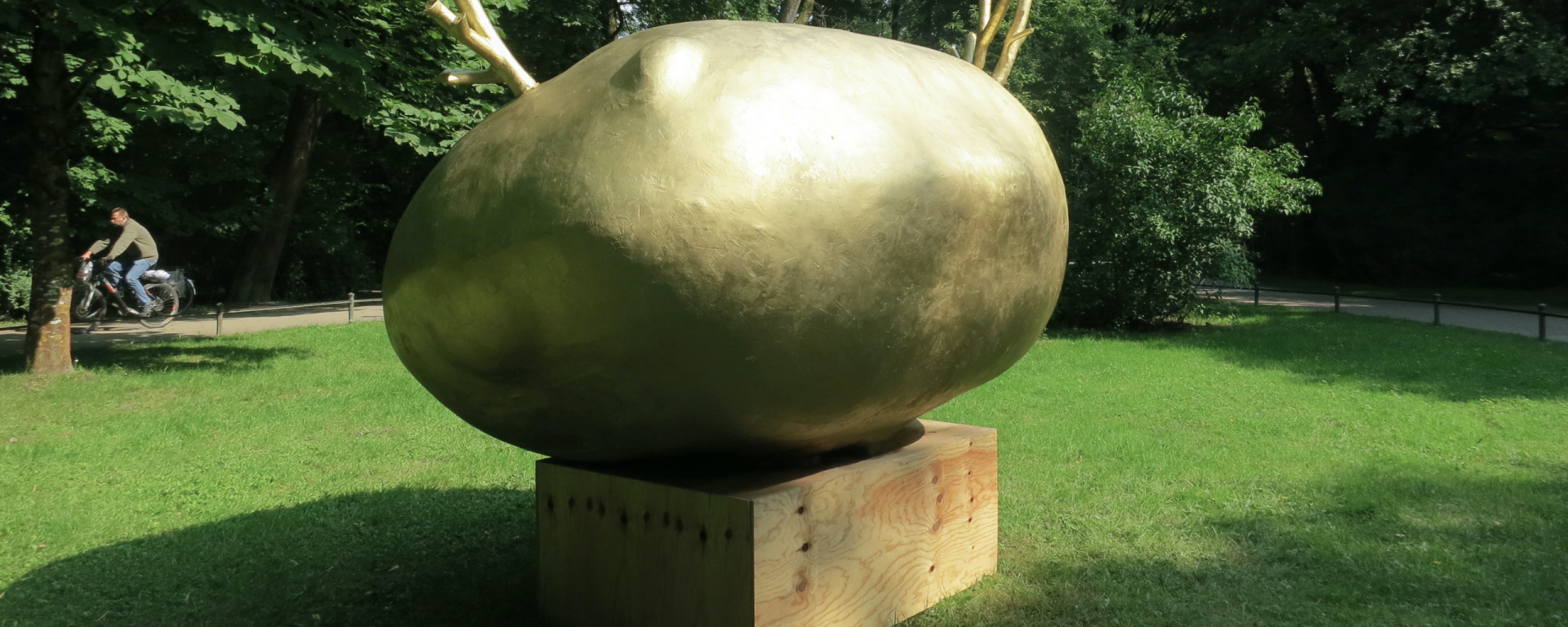 Fotografie von einer Grünfläche im Englischen Garten, auf der eine Installation der Künstlerin Alix Stadtbäumer steht. Die Installation besteht aus einem hölzernen Sockel, auf dem die goldfarbene Skulptur einer Kartoffel ruht, aus der goldene Triebe hervorsprießen.