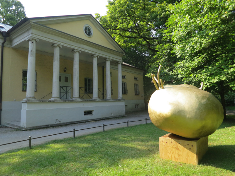 Fotografie von einer Grünfläche im Englischen Garten, auf der eine Installation der Künstlerin Alix Stadtbäumer platziert ist. Die Installation besteht aus einem hölzernen Sockel, auf dem die goldfarbene Skulptur einer Kartoffel ruht, aus der goldene Triebe hervorsprießen. Im Hintergrund zu sehen ist ein klassizistisches Gebäude mit einer Säulenportikus, das sogenannte Rumfordschlössl.