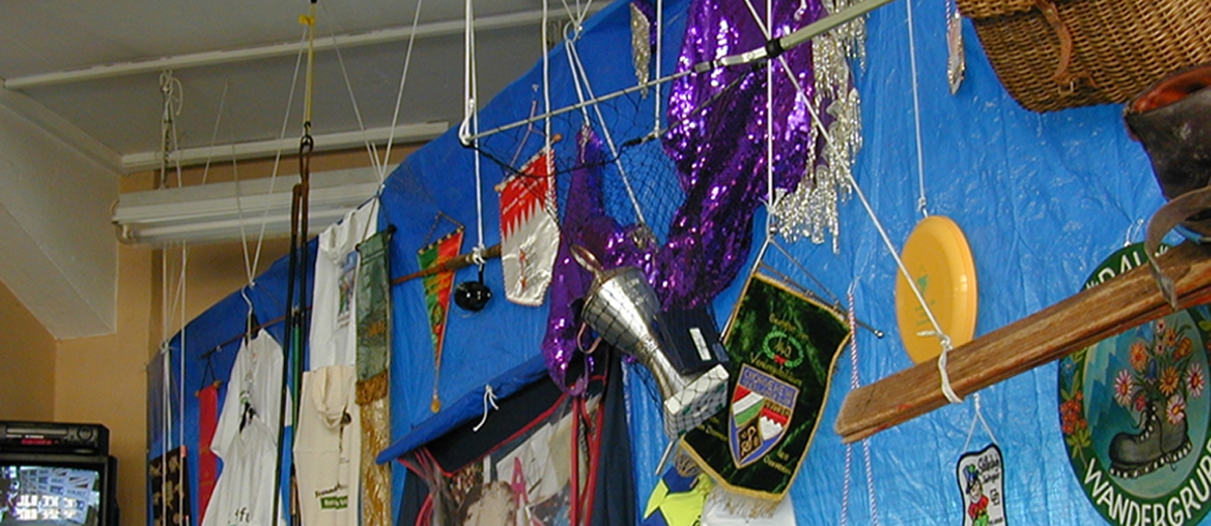 Photograph of a room with a ceiling installation. A large rectangular blue plastic sheet hangs from the ceiling. Various objects from different Munich clubs are attached to this tarpaulin. There are various pennants, t-shirts, baseball caps, trophies, costumes and other club paraphernalia.