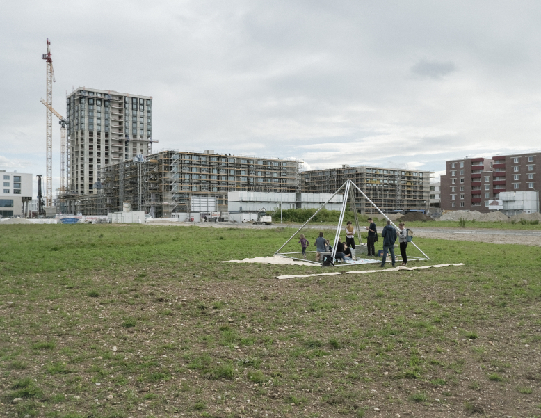 Vor dem Hintergrund einer Großbaustelle mit halbfertigen Hochhäusern steht ein begehbarer Treffpunkt in Form einer Pyramide. Etliche Leute stehen vor und in der Pyramide.