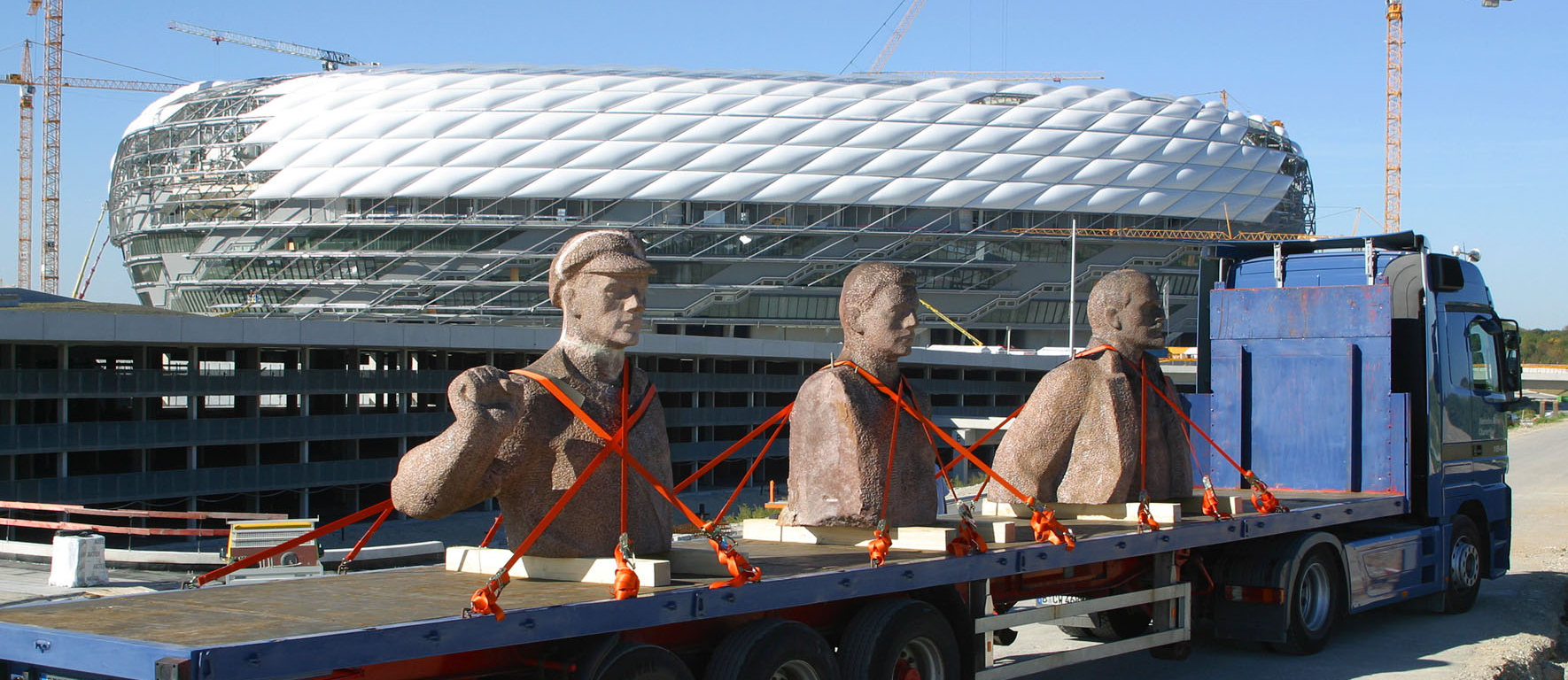 Die Fotografie zeigt im Vordergrund die reisende Skulptur "Lenin on Tour" von Rudolf Herz. Drei Granitbüsten Lenins, die dem Dresdner Lenin-Denkmal entstammen, sind mit orangenen Spanngurten auf einem blauen Sattelschlepper befestigt. Im Hintergrund zu sehen ist die unfertiggestellte, von Kränen umgebene Allianzarena, die sich zu der Zeit im Bau befand.