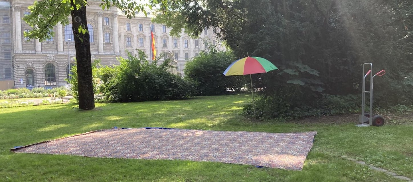 Fotografie einer Grünflache im Alten Botanischen Garten. Auf dem Rasen ist eine große teppichartige Decke ausgelegt, daneben steht eine Sackkarre sowie ein aufgespannter, bunter Sonnenschirm. Im Hintergrund zu sehen ist der Justizpalast.