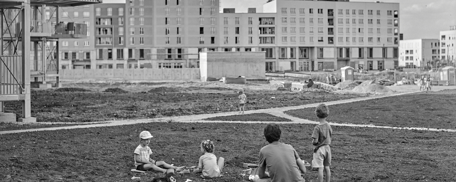 The black-and-white photograph shows several children and an adult sitting or standing in front of Freiham's new buildings; remnants of a building site such as a porta-potty and piles of earth can still be seen
