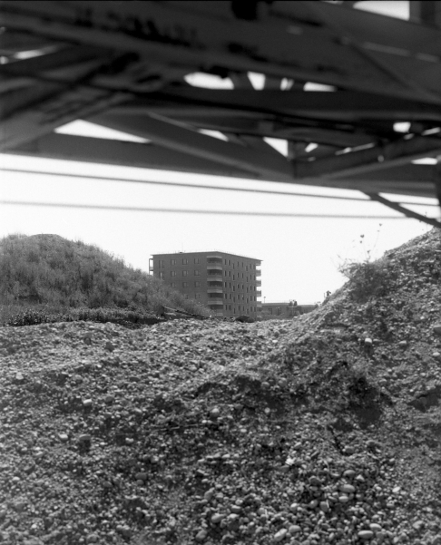 In Schwarz-Weiß zeigt das Bild einen mehrstöckigen Wohnblock in der Ferne, umgeben von Hügeln aus Schutt oder Geröll. Im Vordergrund sind unscharfe Metallstreben und Geröll zu sehen, die den Blick auf das Gebäude teils verdecken.