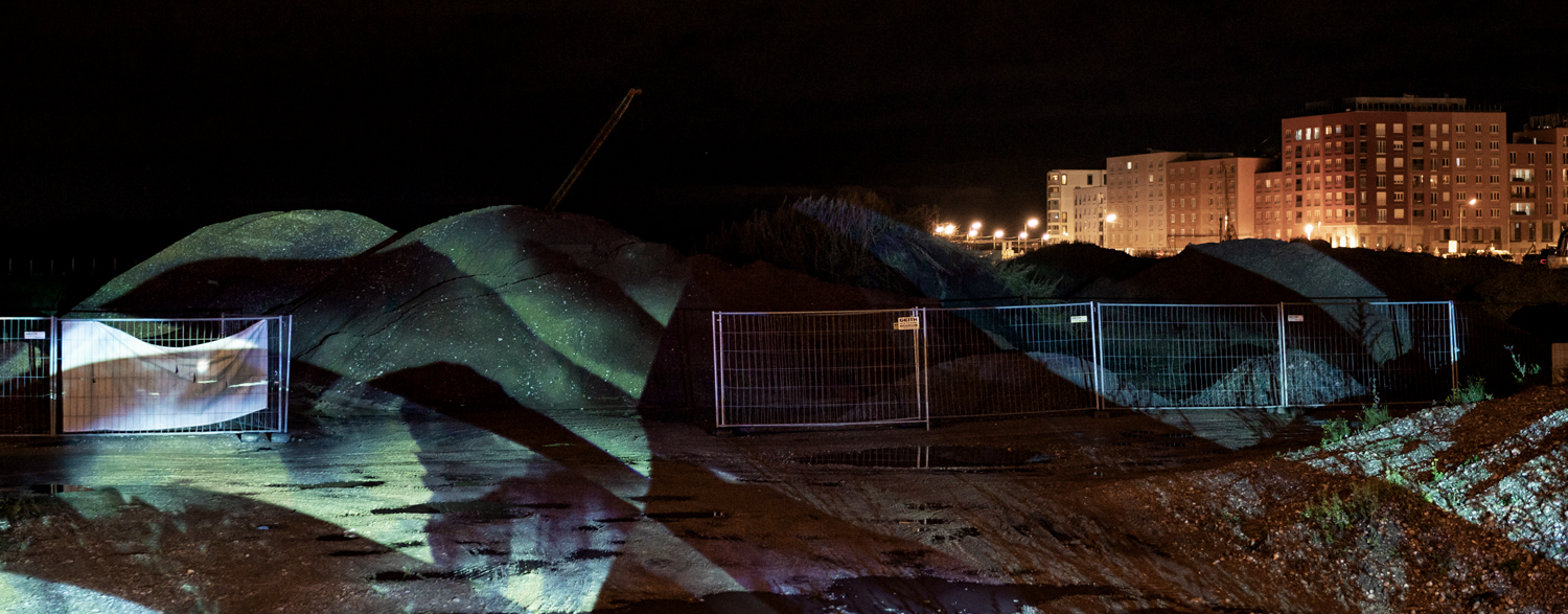 Das Bild zeigt eine Baustelle bei Nacht mit abgesperrten Erdhaufen, auf die eine abstrakte Lichtprojektion geworfen wird. Im Hintergrund sind beleuchtete Wohngebäude zu sehen.