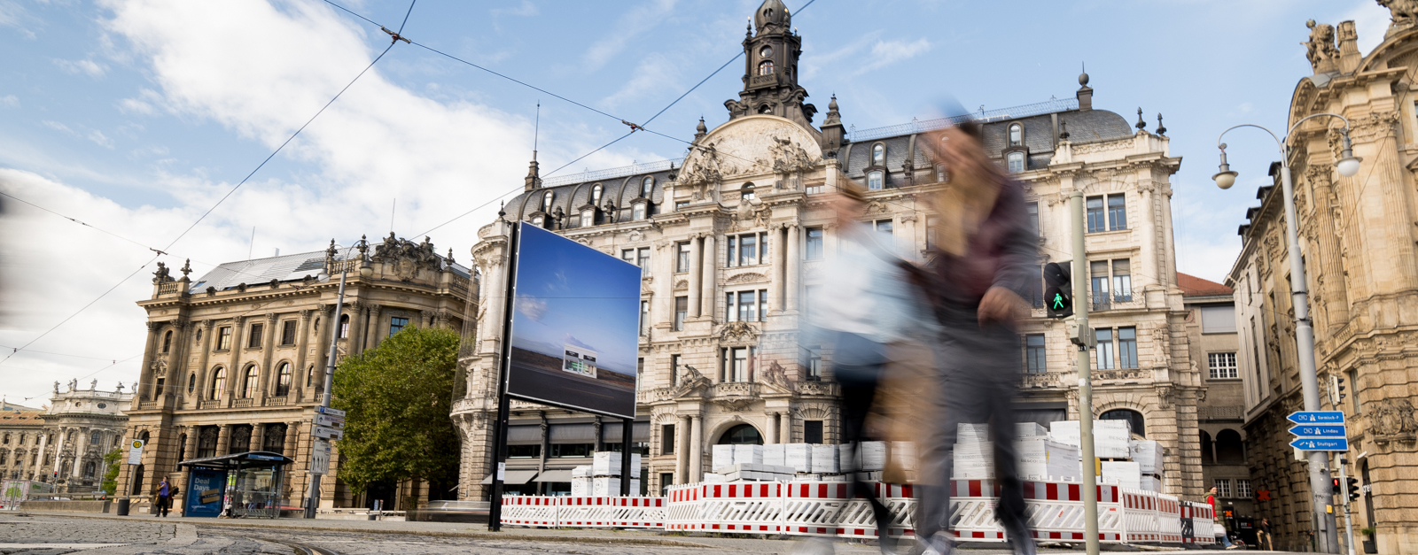 The billboard with the motif by Elmgreen and Dragset shows a lonely Prada boutique in a desert landscape. The billboard stands in front of a historic building in a city, while two people walk past out of focus in the foreground.