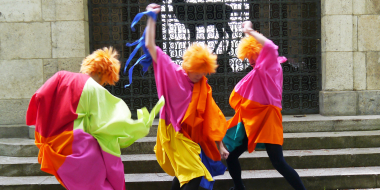 3 Personen mit orangefarbenen Harren in bunten Kostümen bewegen sich wild vor Stufen und einem vergitterten Tor.