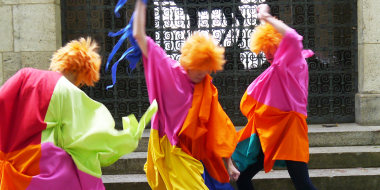 3 people with orange hair in colourful costumes move wildly in front of steps and a barred gate.