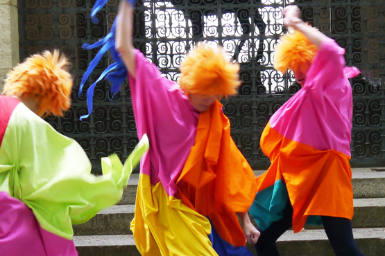 3 people with orange hair in colourful costumes move wildly in front of steps and a barred gate.