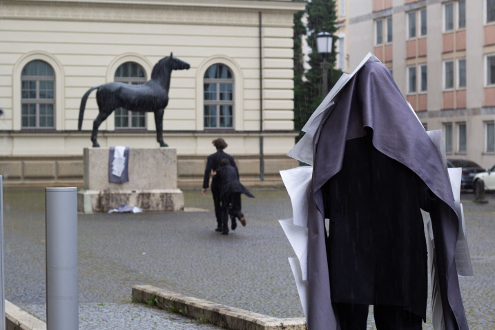 Danke für das Bild! Hier ist ein passender ALT-Text in einfacher Sprache: Eine Person mit einem großen Stoffumhang und daran befestigten Blättern steht im Regen. Im Hintergrund laufen zwei Menschen auf eine Pferde-Statue zu.