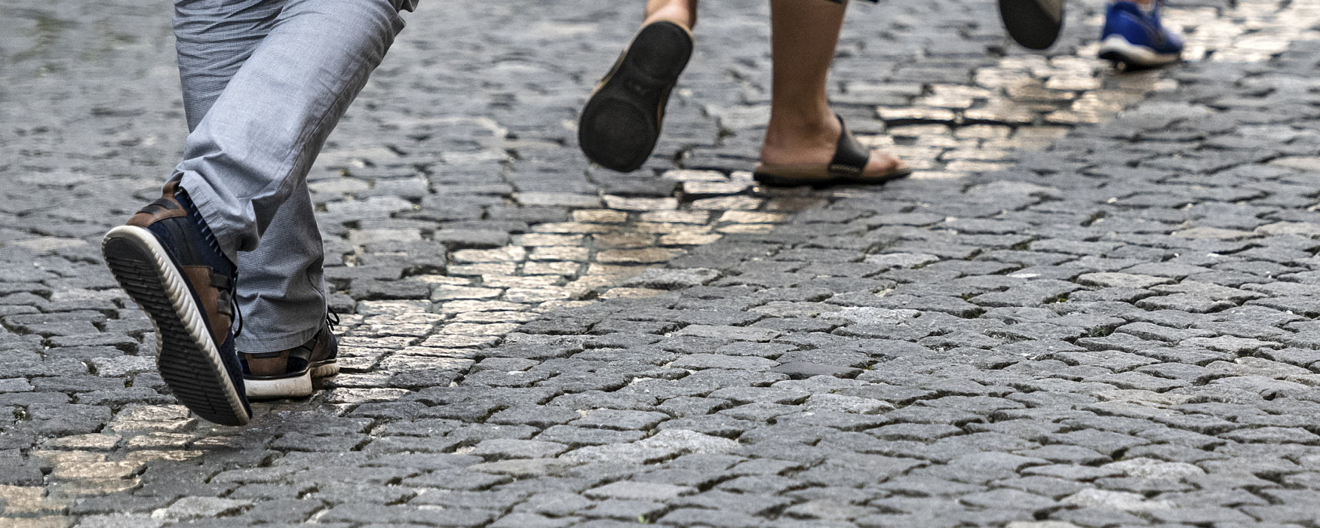 Detailaufnahme einer gepflasterten Fläche, ein Streifen mit einer Breite von ein bis vier Steinen hebt sich durch eine gold-glänzende Oberfläche ab, dieser Streifen wird von drei Personen beschritten, von denen fast nur die Füße zu sehen sind.