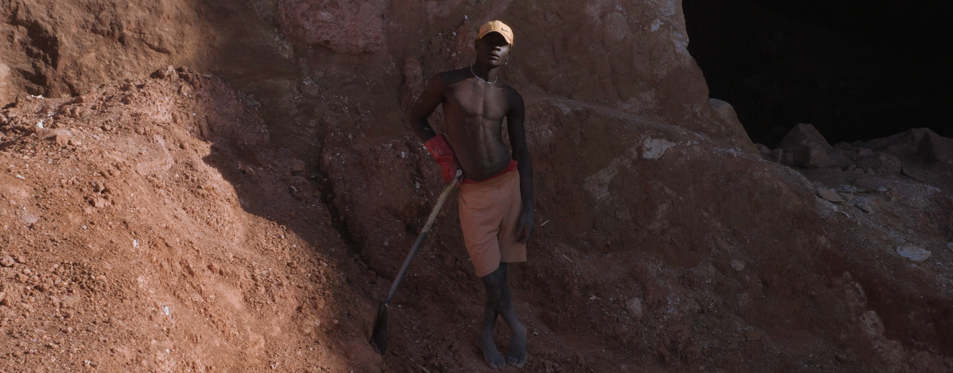 Person stands with naked upper body in front of a reddish rock face, leaning on a shovel.