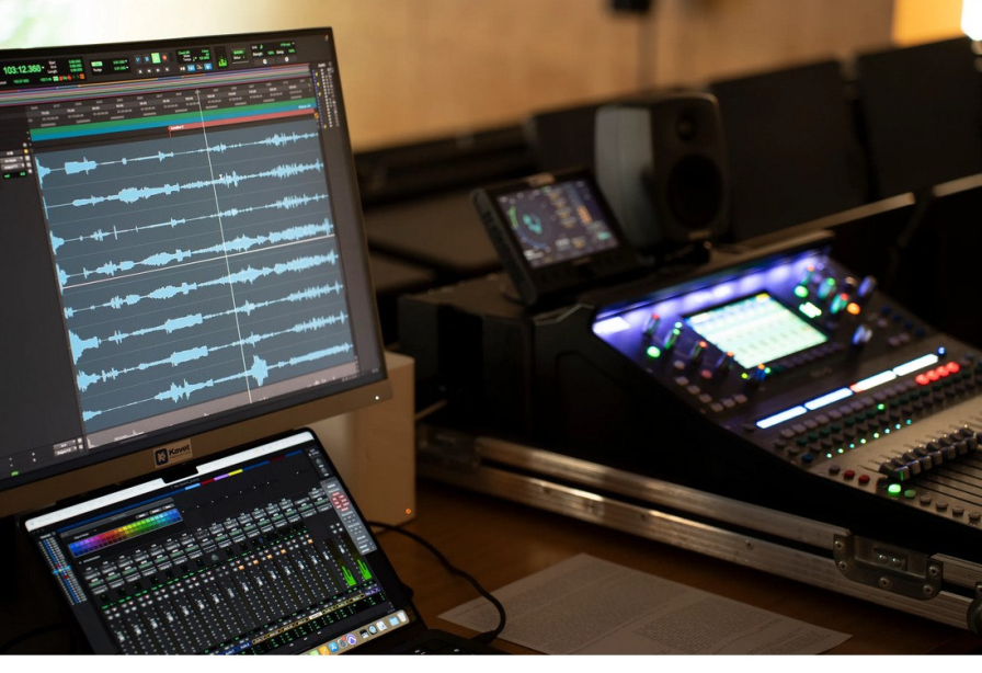 Blick von schräg oben auf ein Mischpult und ein Notebook sowie ein Monitor, darauf zu sehen verschiedene Tonspuren und Equalizer.
