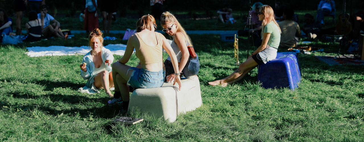 zwei bankähnliche Objekte in beige und lila, stehen im Gras, sommerlich bekleidete Personen sitzen darauf bzw. auf dem Boden, im Hintergund liegen Decken.