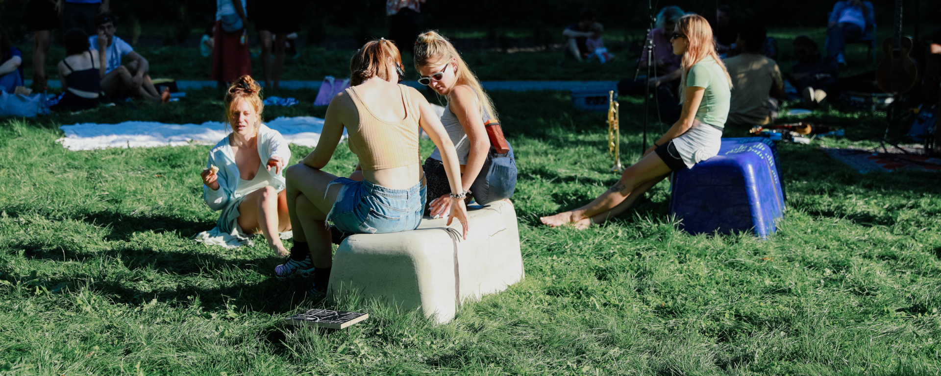 zwei bankähnliche Objekte in beige und lila, stehen im Gras, sommerlich bekleidete Personen sitzen darauf bzw. auf dem Boden, im Hintergund liegen Decken.