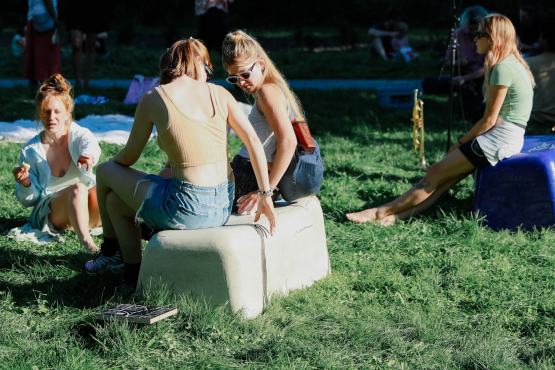 zwei bankähnliche Objekte in beige und lila, stehen im Gras, sommerlich bekleidete Personen sitzen darauf bzw. auf dem Boden, im Hintergund liegen Decken.