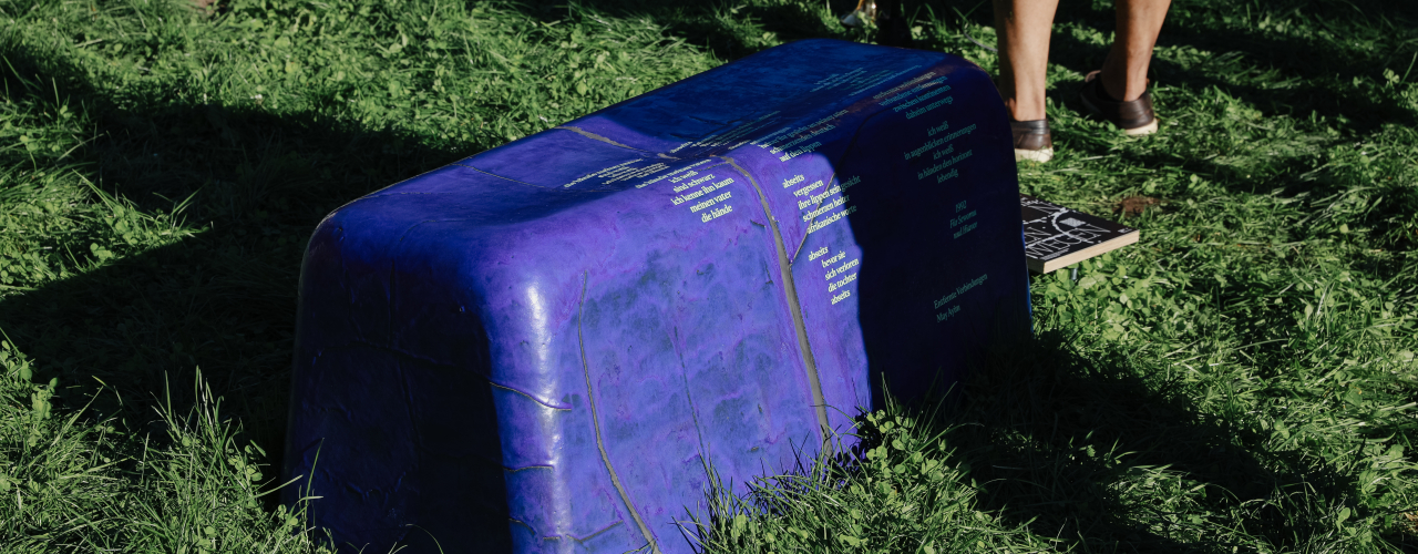 Lila bankähnliches Objekt mit gelblicher Schrift steht im Gras, daneben Beine von Personen.