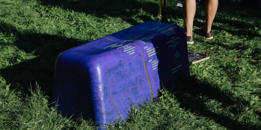 Lila bankähnliches Objekt mit gelblicher Schrift steht im Gras, daneben Beine von Personen.