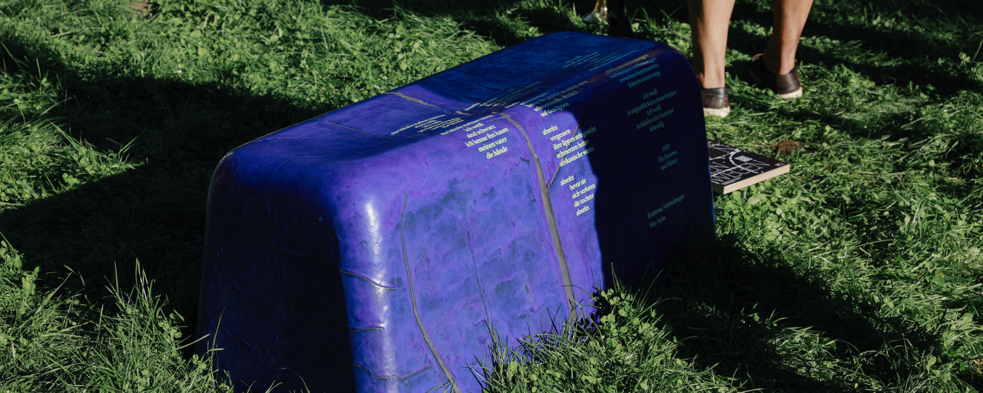 Lila bankähnliches Objekt mit gelblicher Schrift steht im Gras, daneben Beine von Personen.