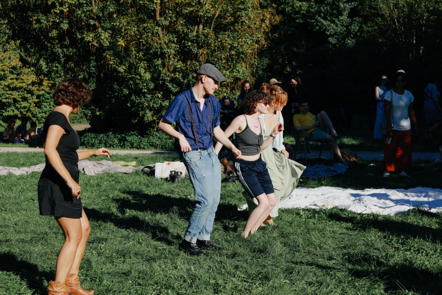 Mehrere sommerlich gekleidete Personen tanzen teils barfuss im Gras, im Hintergrund liegen Decken.