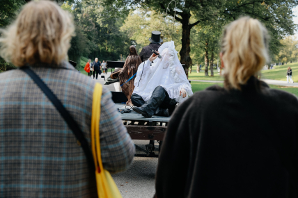Eine Kutsche fährt durch einen Park, auf der Kutsche steht eine bräunliche Madonnenskulptur und sitzt eine von weißer Spitze verhüllte Person, die ein Mikrofon in der Hand hält.