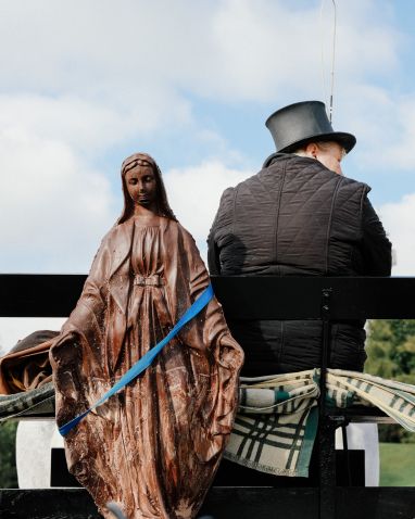 Frontalansicht einer großen Madonnenskulptur aus braunem Material vor leicht bewölktem Himmel, dahinter sitzend eine schwarz gekleidete Person mit Zylinder.