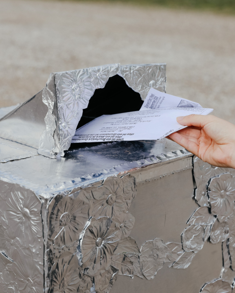 Eine Hand steckt einen Brief in einen Briefkasten, der mit silberner, blümchengeprägter Folie überzogen ist.