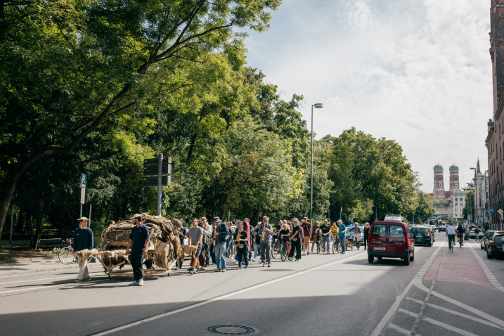 Several people push an artistic object, the remains of a crumbling SUV made of clay, through a street, followed by a few people. The towers of Munich's Frauenkirche can be seen in the background.