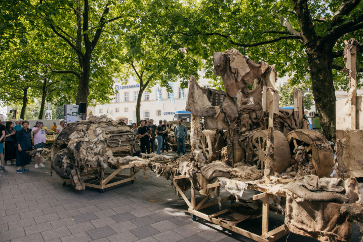 The remains of the crumbling SUVs made of clay on wooden scaffolding stand in a square under trees, with a few people standing around them.