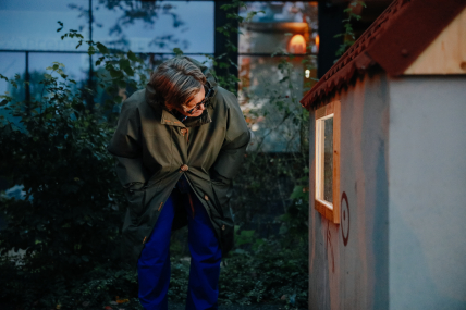 Person beugt sich in der Dämemrung zu einem kleinen Haus und schaut ins Fenster.