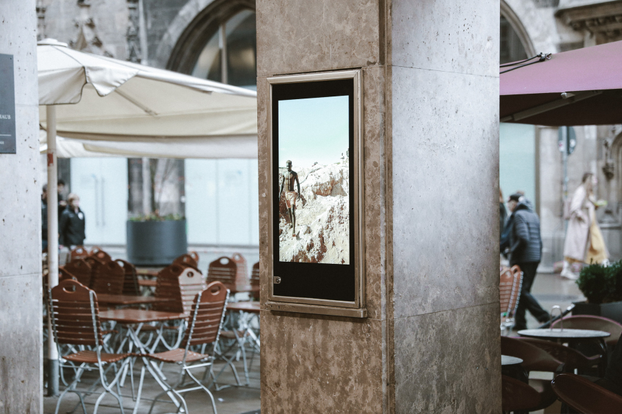 Vitrine an einem Pfeiler mit einem digitalen Bildschirm, auf diesem ist eine Person in einer Felslandschaft zu sehen. Im Hintergrund leere Stühle eines Straßencafés.