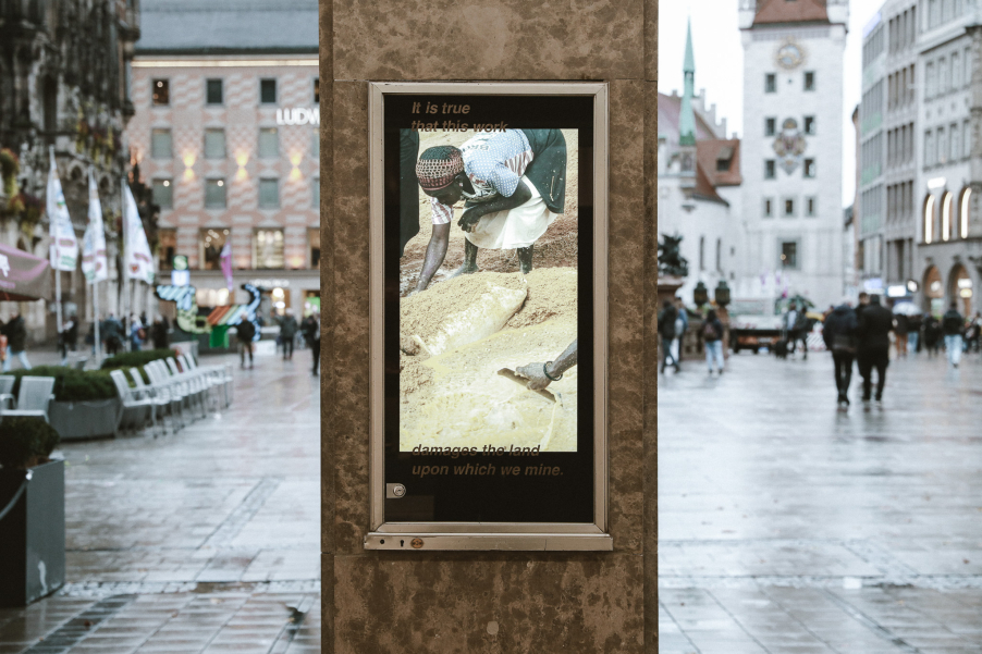 Vitrine an einem Pfeiler mit einem digitalen Bildschirm, auf diesem ist eine barfüßige Person gebückt im Schlamm stehend zu sehen. Im Hintergrund öffentlicher Straßenraum.