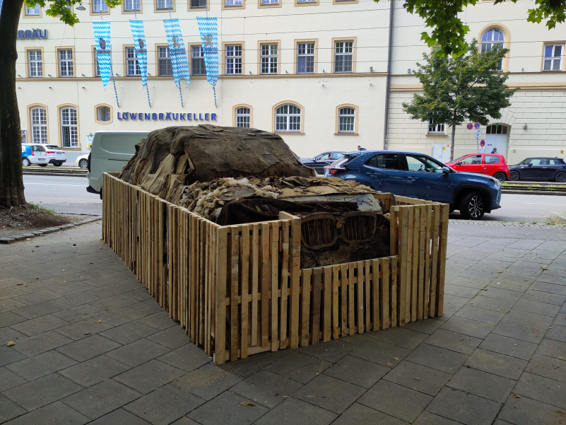 The crumbling SUV made of clay is fenced in with a wooden fence, in the background you can see a large building with the words 'Löwenbräukeller' on it