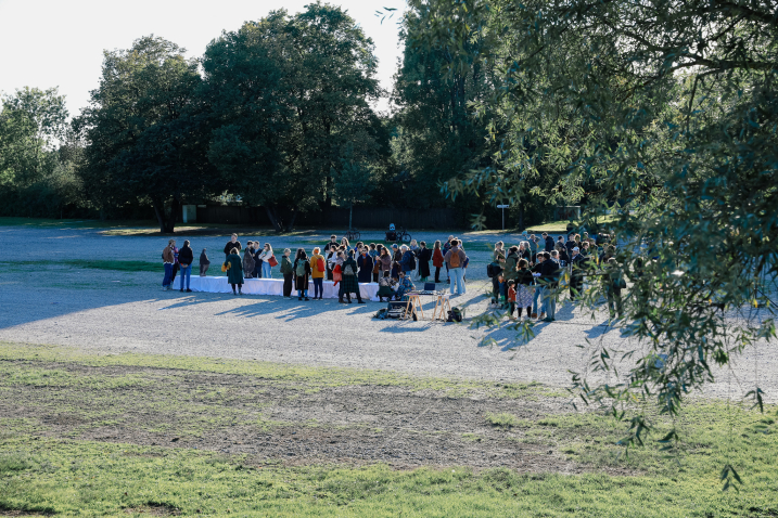 Im Freien stehen viele Menschen auf einer Schotterfläche um eine lange Tafel herum, die mit weißen Tüchern bedeckt ist. Im Hintergrund Bäume.