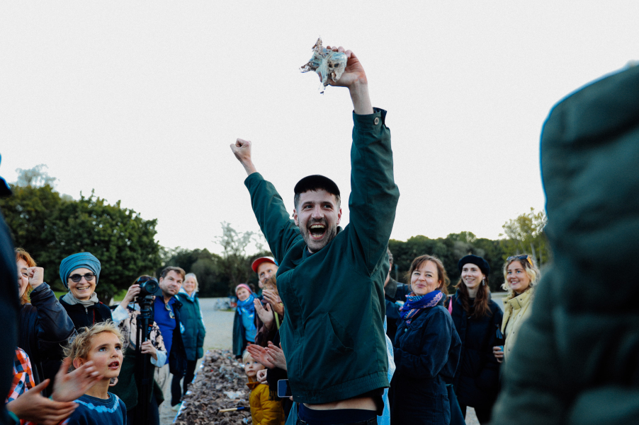 Person hält jubelnd ein zerbrochenes Fragment in die Luft, umringt von applaudierenden und lachenden Menschen im Freien.