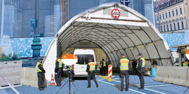 Eine temporäre Kontrollstelle mit Zeltdach und mehreren Sicherheitskräften steht auf einem Platz vor einem historischen Gebäude mit Gerüst und Kran.