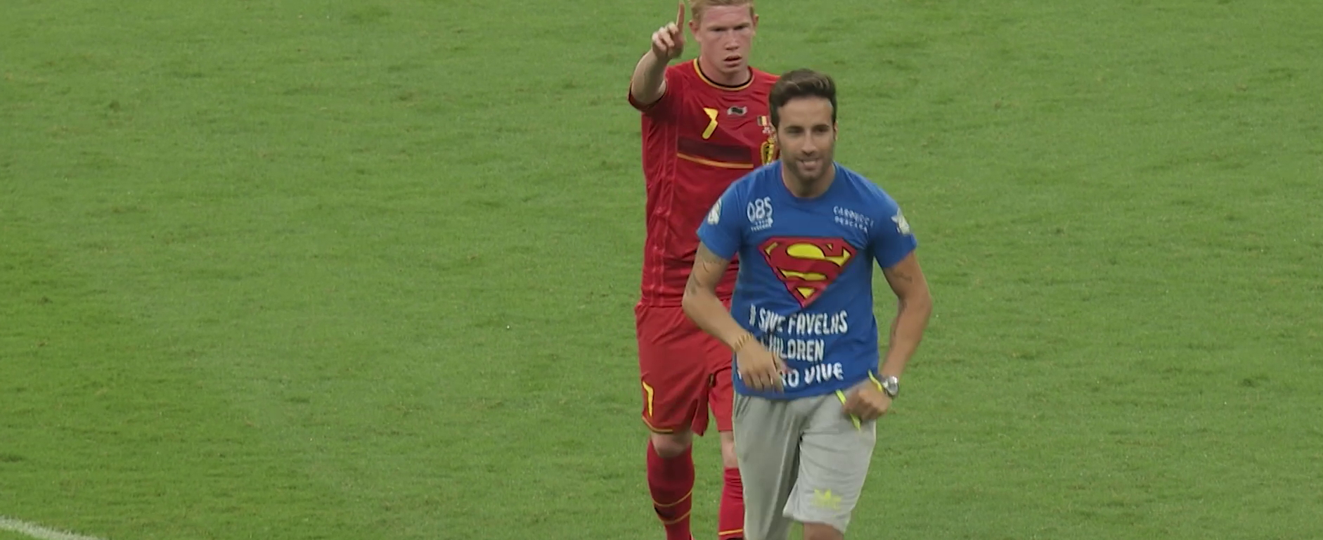 Soccer player in a red jersey points forward, in front of him a man in a blue T-shirt runs across the grass.