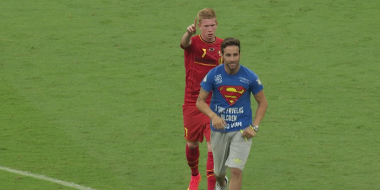 Fußballspieler im roten Trikot zeigt mit dem Finger nach vorn, vor ihm läuft ein Mann im blauen T-Shirt über den Rasen.
