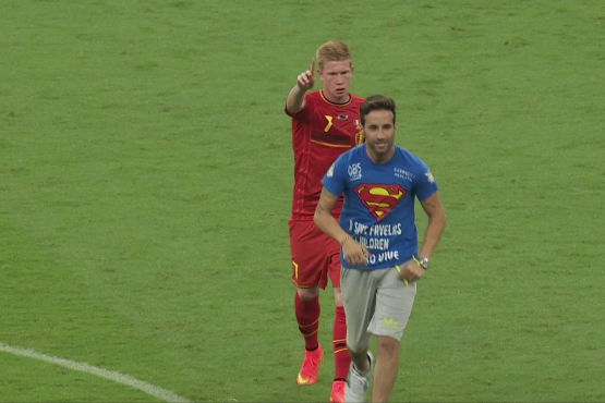 Fußballspieler im roten Trikot zeigt mit dem Finger nach vorn, vor ihm läuft ein Mann im blauen T-Shirt über den Rasen.