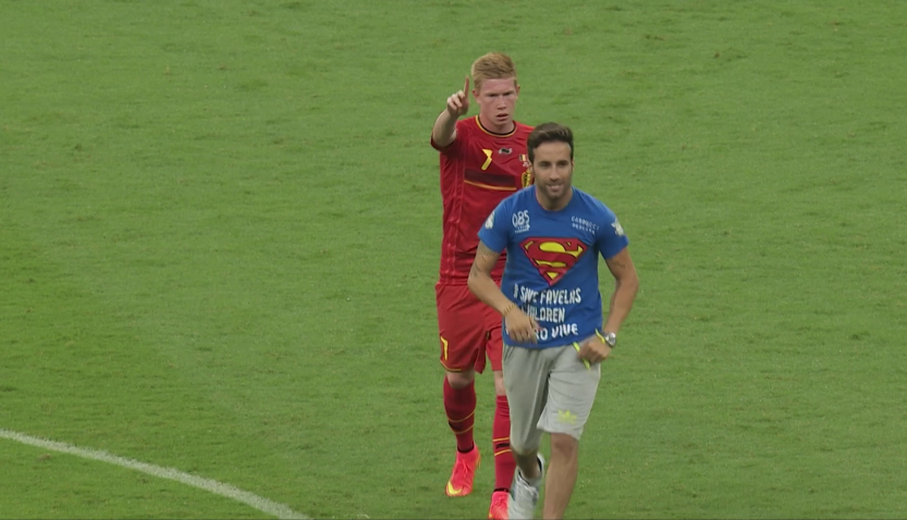 Fußballspieler im roten Trikot zeigt mit dem Finger nach vorn, vor ihm läuft ein Mann im blauen T-Shirt über den Rasen.