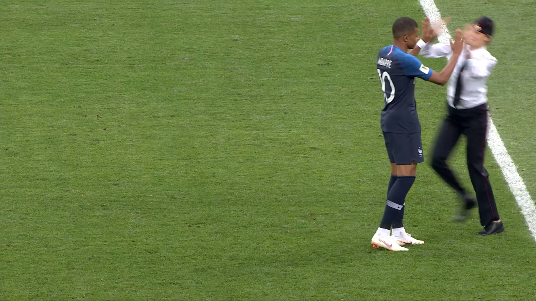 Eine Person in dunkelblauem Fussballtrikot steht auf einem Fussballfeld und klatscht mit einer Person in schwarzer Hose, weißem Hemd und schwarzer Krawatte ab.