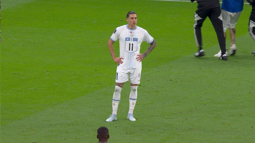 Frontalansicht eines Flussballspielers auf dem Fussballfeld mit der Nr. 11 in weißem Trikot. Rechts oben im Hintergrund die Beine von drei Personen, der Kleidung nach offensichtlich keine Spieler*innen.